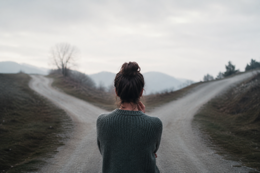 Mulher parada diante de uma bifurcação de caminhos, observando as duas direções com expressão reflexiva e contemplativa, simbolizando a dúvida sobre ter tomado a decisão certa após uma mudança importante na vida.