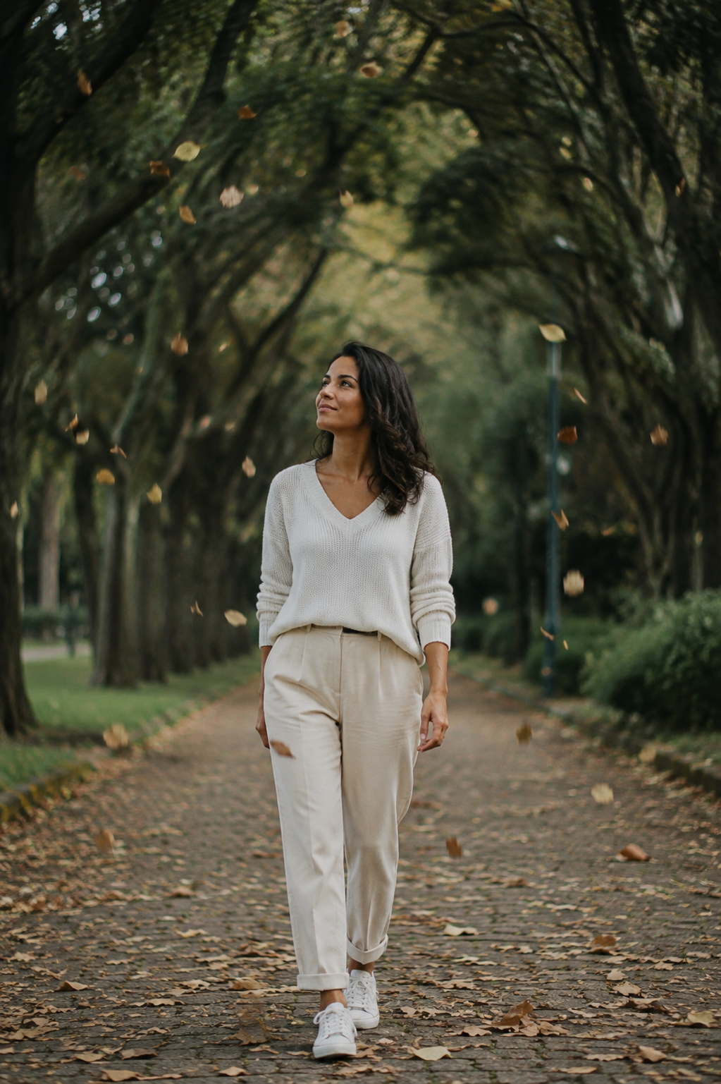 Mulher parda caminhando lentamente por um jardim arborizado enquanto folhas caem ao seu redor, com expressão serena e esperançosa, simbolizando a transformação da saudade e a capacidade de seguir em frente após um término de relacionamento.